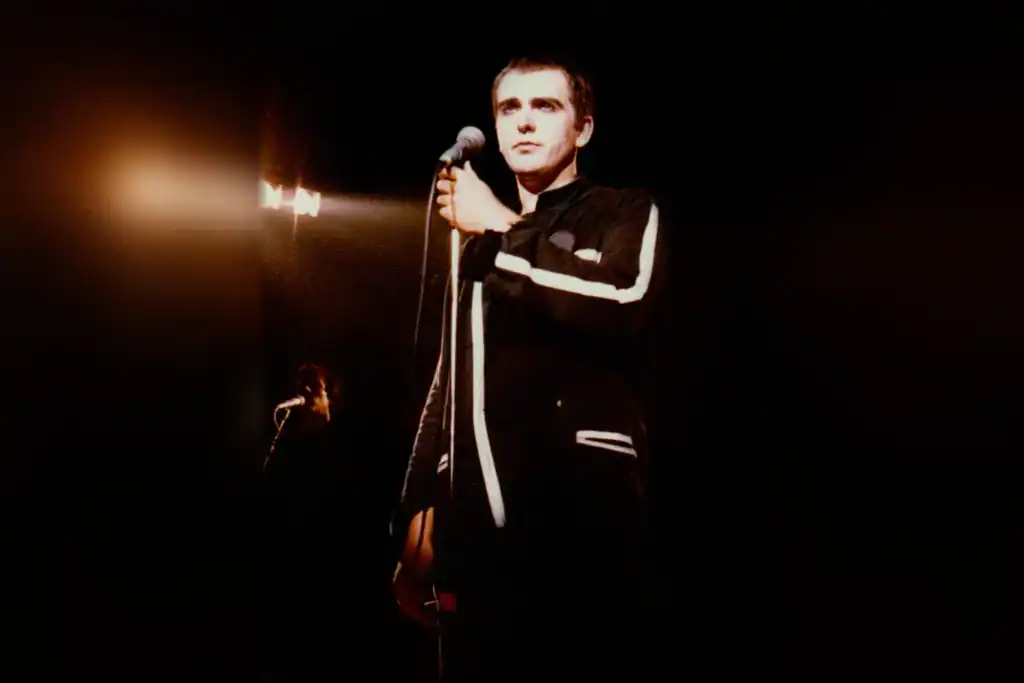 Peter Gabriel holding a microphone on stage, wearing a black jacket with white trim.